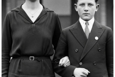 A Mother and Her Son in 1925 — Experts Analyze the Photo and Are Left Speechless  A mother and her son in 1925.  Experts analyzed the photo and are left speechless.  Dr.Sarah Mitchell adjusted her reading glasses as she carefully examined the contents of the donated estate collection at the Smithsonian’s National Museum of American History.  Among the dusty boxes of documents and memorabilia from the Henderson family of Chicago, one particular photograph caught her attention.  The black and white image dated 1925 showed a well-dressed woman in her 30s standing beside a boy of perhaps 12 years old.  They posed formerly in front of what appeared to be a modest Chicago brownstone, both wearing their finest clothes.  What struck Sarah immediately wasn’t the subjects themselves, but something peculiar in the background.  Behind the mother’s left shoulder, barely visible in the window of the house, was a reflection that seemed oddly out of place for the era.  She reached for her magnifying glass.  her heart beginning to race as she focused on the mysterious detail.  The image quality was remarkably sharp for a 1925 photograph.